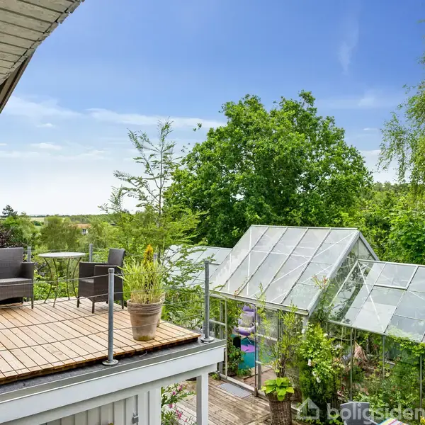 En træterrasse med stol og bord har udsigt over en have med et stort drivhus. Omkring drivhuset er der frodig grøn vegetation og en klar blå himmel.