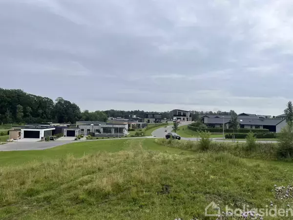 Flere moderne huse står langs en stille vej i et grønt, let bakket landskab med skov baggrunden under en overskyet himmel.