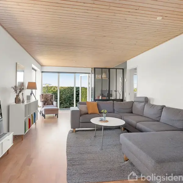 Stor grå hjørnesofa står i en moderne stue med bræddegulve og loft. Et sofabord står foran sofaen. I baggrunden er store vinduer, der slipper lys ind, og en reol ved væggen.