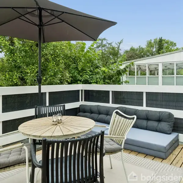 Terrasse med rundt bord og fire stole under en stor parasol, omringet af træhegn. En blå sofa med hynder står ved siden af. Grønt løv og tag ses bagved.
