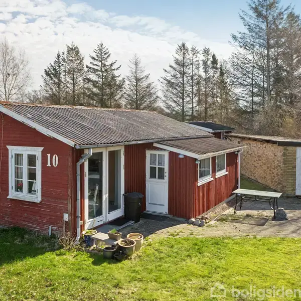 Rødt hus med nummeret "10" på væggen står på en græsplæne. Ved siden af er en garage, og der er træer i baggrunden. Potteplanter og et bord ses foran.
