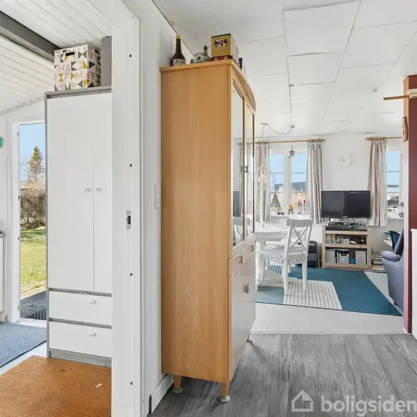 En entré med et klædeskab står ved siden af en træskab. Foran ses en stue med spisebord og sofa, og i baggrunden er en græsplæne synlig gennem vinduerne.