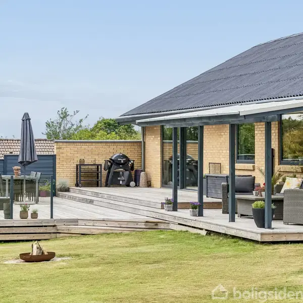 Et moderne hus med en overdækket terrasse, indeholder havemøbler og planter. Terrassen har udsigt over en græsplæne, og der er en grill samt et fritstående bålfad i haven.