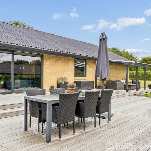 Et moderne træbord med sorte kurvestole står på en stor træterrasse foran et gult murstenshus. Der er en stor parasol ved bordet, og omgivelserne inkluderer grønne træer og en græsplæne.