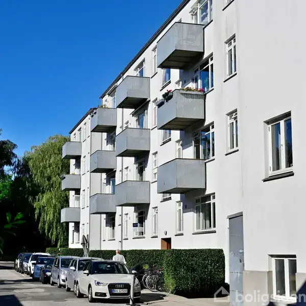 En hvid, fler-etagers bygning har flere altaner og er omkranset af en stille gade med parkerede biler og træer i baggrunden. En person går langs fortovet.