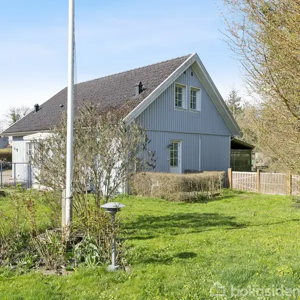 Et gråmalet hus med skråt tag står i en grøn have med træer og buske. En flagstang er placeret i forgrunden, og huset har en indhegnet terrasse.