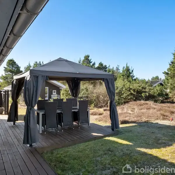 Pavillon med spisebord og stole står på en terrasse ved siden af et hus. Omgivet af en naturgrund med buske og træer under en klar blå himmel.