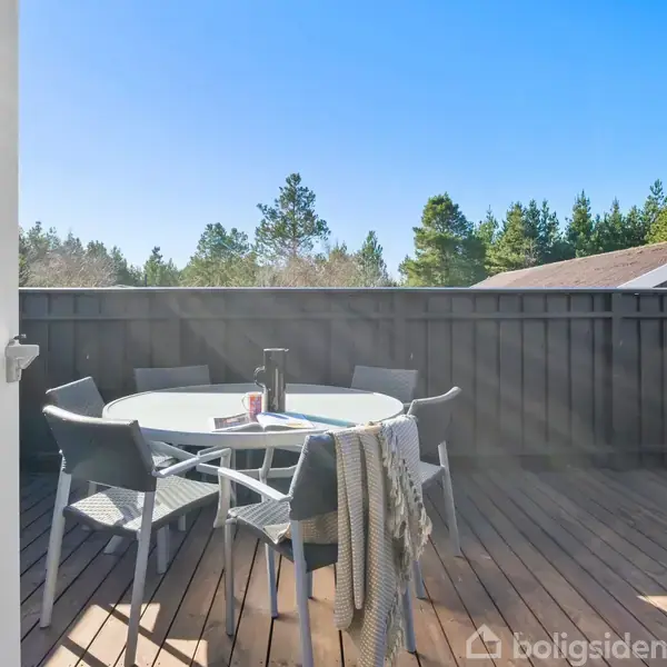 En rund terrassebord med stole står solbeskinnet på en træterrasse. En kop og en bog ligger på bordet. Omgivet af et højt træhegn og grønne træer.