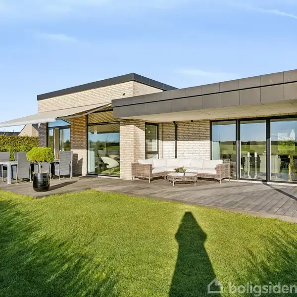 Et moderne hus med store glasfacader står på en grøn græsplæne. Haven har en terrasse med havemøbler, og en blå himmel ses i baggrunden.