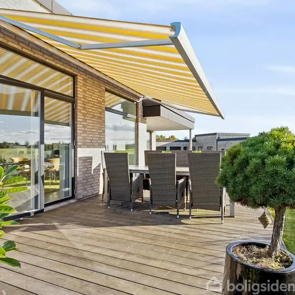 En terrasse med trædæk har en spisebordssæt under en udtrukket gul-og-hvid-stribet markise. Omkranset af planter og udsigt over en åben mark og bygninger i det fjerne.