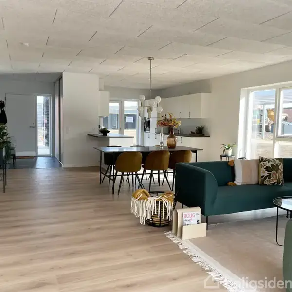 Stue med moderne spisestue, sofagruppe og grøn sofa. Store vinduer lader lys ind. Blomster og bøger dekorerer rummet. Rundt spisebord med stole i træ og en lys lampe hængende over.