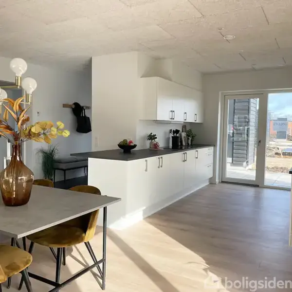 Et stilfuldt køkken med hvide skabe og en mørk bordplade. Et spisebord med stole står i forgrunden, omgivet af en stor vinduesdør, der fører ud til en terrasse.