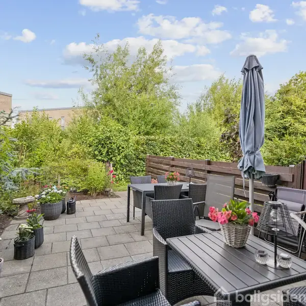 Terrasse med sorte kurvemøbler arrangeret ved et bord, omgivet af blomster i urtepotter. Parasol står ved siden af møblerne. Baggrunden har tæt grøn beplantning og en klar blå himmel.