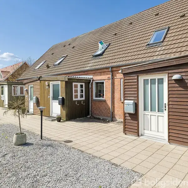 Rækkehus med brun facade, hvide døre og vinduer står under en klar blå himmel. Forhaven har fliser og grå grus, omgivet af buske og træer i baggrunden.