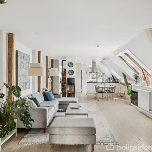 En lys stue med en grå sofa og grønne planter står ved siden af et spisebord i baggrunden. Skråvinduerne lukker naturligt lys ind, og dekorative elementer ses langs væggen.