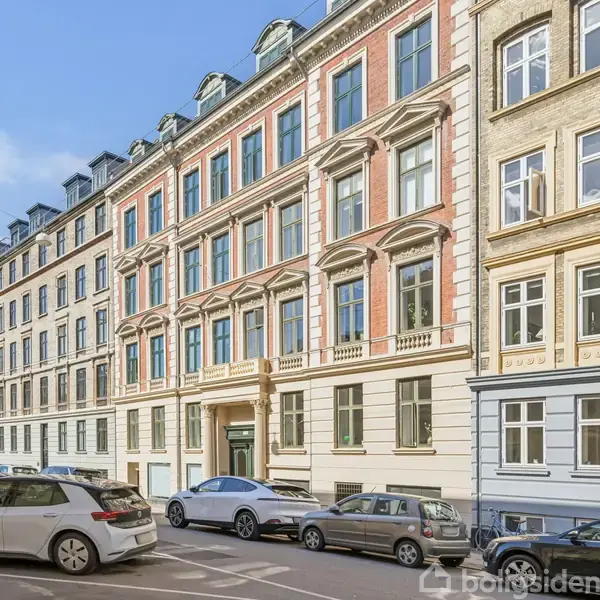 Flere etageejendomme står langs en byvej. Biler er parkeret på begge sider af vejen. Bygningerne har klassiske arkitektoniske træk og flere vinduer.