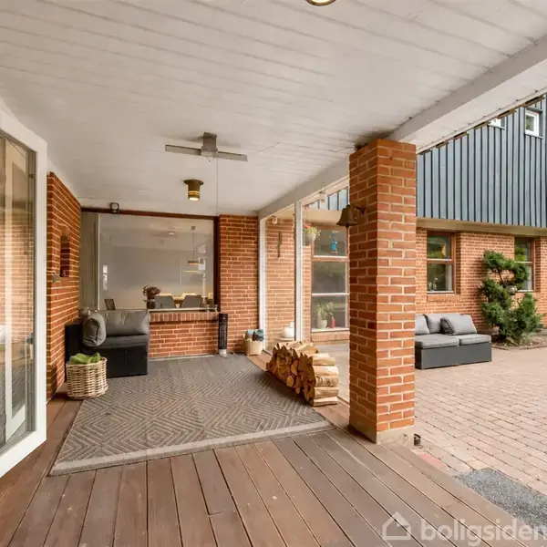 Terrasse med trædæk og murstensstolper, der åbner op til et moderne boligområde. Møbler som sofa og planter skaber en hyggelig atmosfære. Åben dør leder ind til huset.