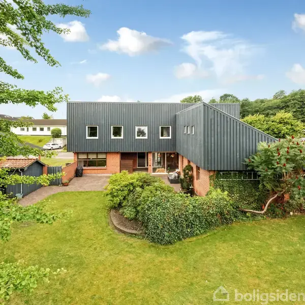 Et moderne hus med fladt tag står i en velplejet have med græsplæne og blomsterbede. Omkringliggende grønne områder og nabohuse er synlige i baggrunden under en blå himmel med spredte skyer.