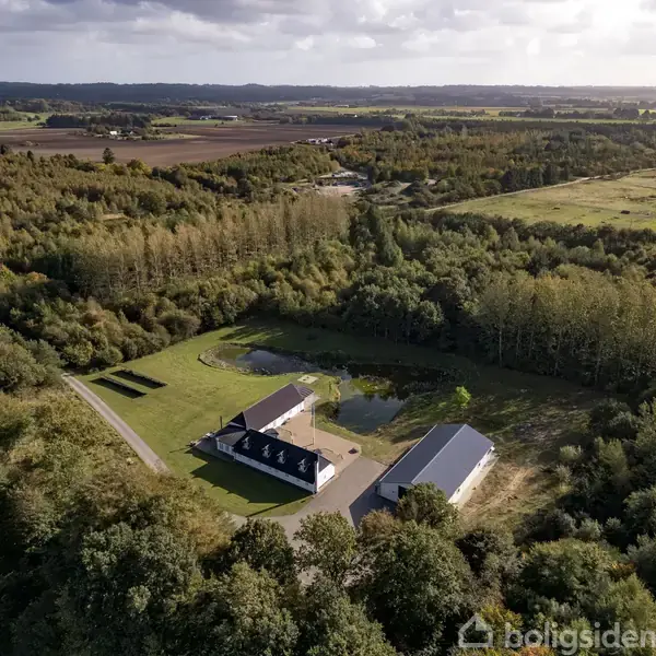 Hus og lade ligger i en skovklædt landskab med åbne marker i baggrunden under en blå himmel. En vej fører til ejendommen, der er omgivet af træer og en dam.