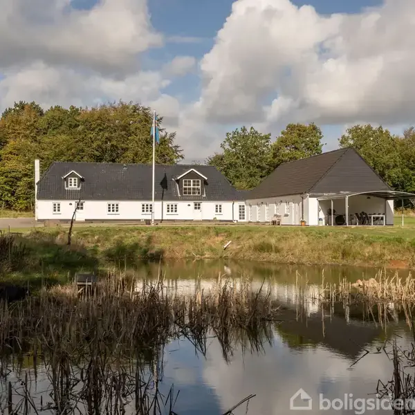 En hvid bygning med sort tag ligger ved siden af en dam omgivet af græs og træer under en delvist skyet himmel.
