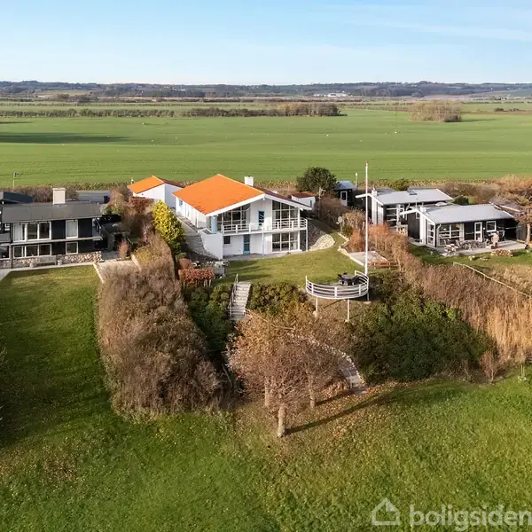 Hvidt hus med orange tag står på en højtliggende grund med udsigt over en stor, åben mark. Omkringliggende huse og beplantning skaber en fredelig, naturskøn atmosfære.