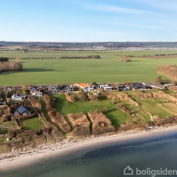 Huse ligger fredeligt langs en kystlinje, med frodige grønne marker i baggrunden. Kystens sand og blide bølger danner en harmonisk overgang mellem land og vand.