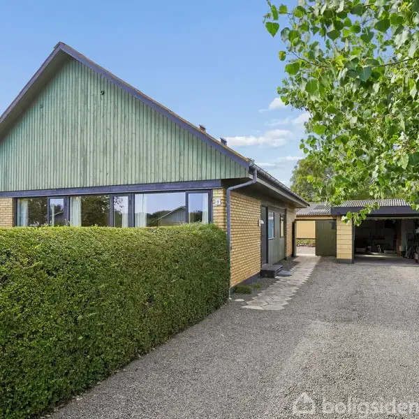 Hus med grønt tag, omgivet af tætklippede hække, står stille på en grusvej ved siden af en garage og frodig vegetation.