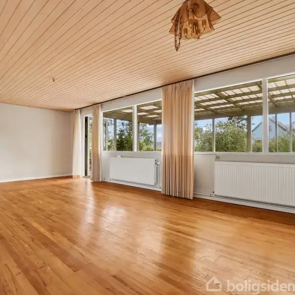 Et tomt rum med trægulv og lyst træloft, store vinduer med udsigt til grønne omgivelser. To radiatorer placeret under vinduerne, og gardiner hænger ved siden af dem.