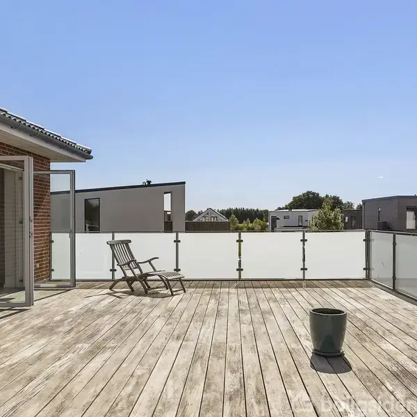 En stor træterrasse med to tomme plantepotter og en liggestol, i sollys, omgivet af glasrækværk og udsigt til moderne huse og blå himmel.