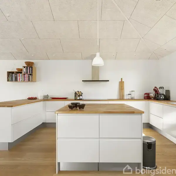 Et lyst køkken med moderne, hvide skabe. En stor køkkenø står centralt. Maleri og reoler hænger på væggen. Rummet har træarbejdsflader og en pendellampe over øen.