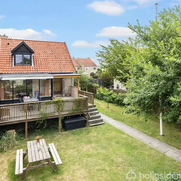 Rækkehus med rødt tegltag og træterrasse; terrasse har siddepladser. Græsplæne med picnicbord i haven, omgivet af grønne træer og buske. Solrig dag med blå himmel.