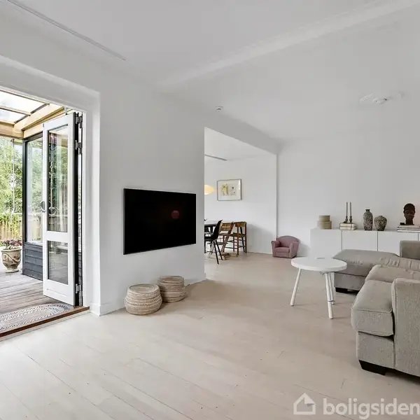 Stue med grå sofa, sofabord og dekorationer på skænken. Åben dør fører til terrasse med haveudsigt. Lysindfald fra vinduerne skaber en lys og rummelig atmosfære.