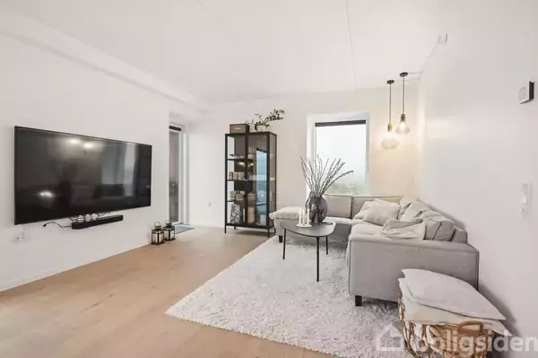 En grå sofa står mod en væg med hængende lamper ved siden af et rundt sofabord med en vase på. En reol er placeret i rummet, der har lyse trægulve. En stor fladskærms-TV hænger på en hvid væg.