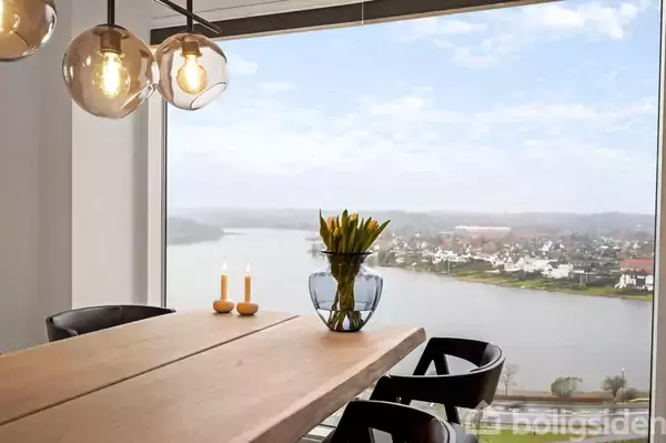 En spisebord med blomster i en vase står ved et vindue med udsigt over en sø og huse. To tændte lys er på bordet, og loftslamper hænger over.
