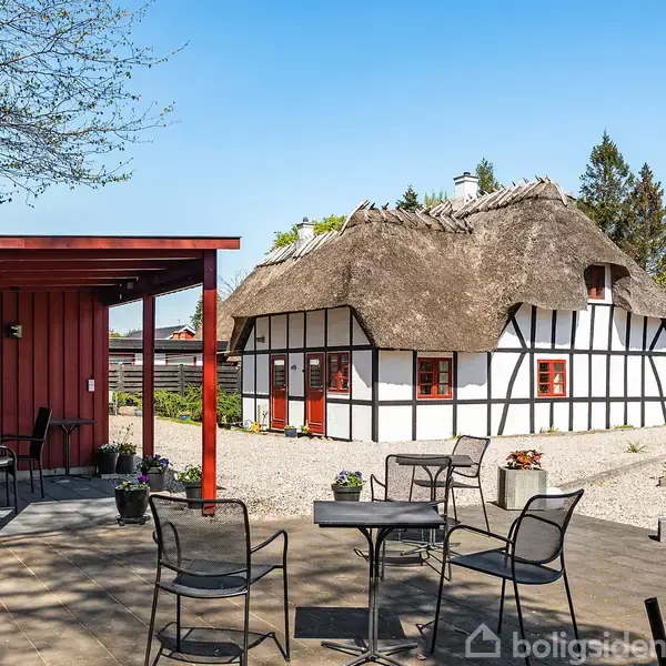 Bindingsværkshus med stråtag står stille i en have. Foran er en terrasse med borde og stole. Omkring er grønne træer og bede med blomster.