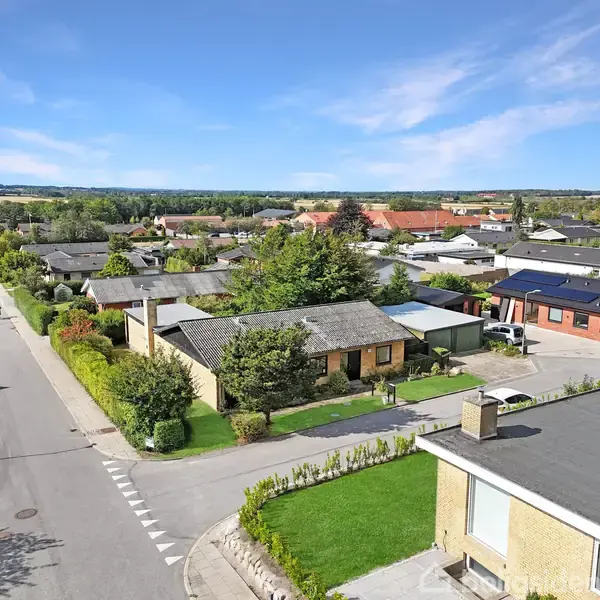 Et parcelhus med fladt tag står omgivet af grønne haver i en rolig kvartervej. Flere lignende huse og træer ses i baggrunden under en klar blå himmel.