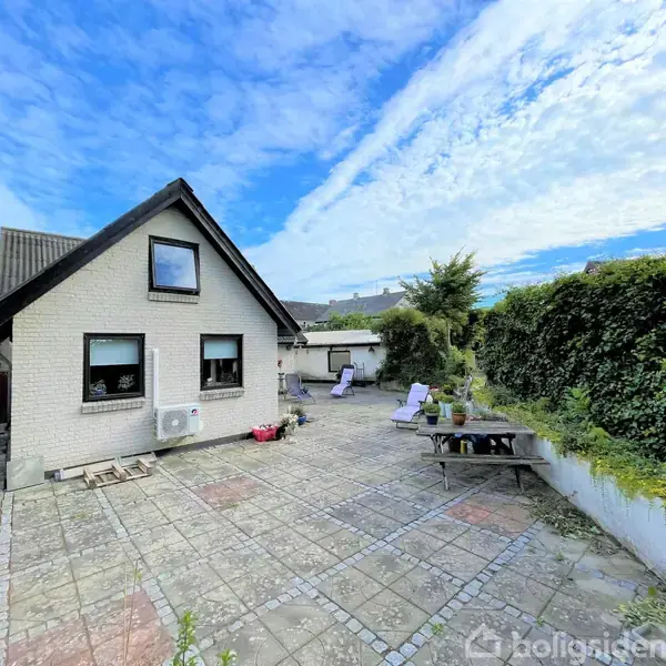 Et hvidt hus med en sort skråtag står i en flisebelagt gårdhave med havemøbler og planter. Baggrunden viser en klar blå himmel og buske langs højre side.