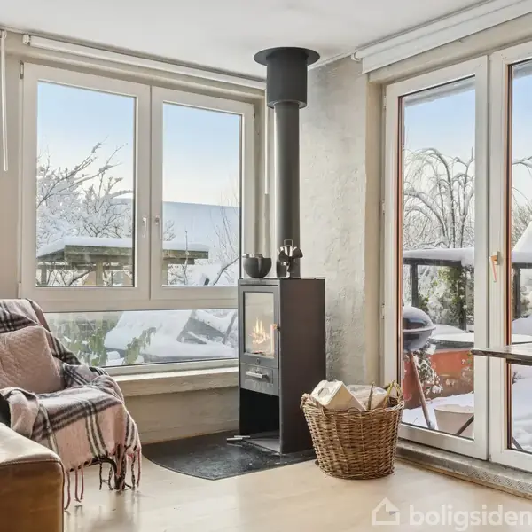 En brændeovn står tændt i et opholdsområde med store vinduer, der afslører en snedækket baghave. En lænestol med tæppe står ved siden af en sofa, der skaber en hyggelig atmosfære.
