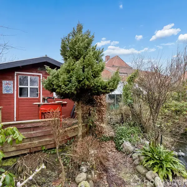 En rød træhytte står stille ved en dam omgivet af buske og træer i en have. Ved siden af hytten er et skilt og en lille udendørsbænk.