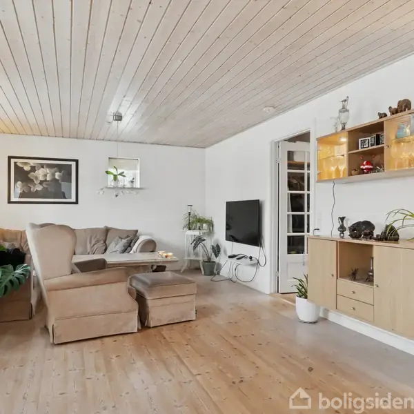 Stue med beige sofa og lænestol indrettet med grønne planter. En reol med dekorative genstande hænger på væggen. Hvidmalede vægge og lofter samt trægulv skaber en lys atmosfære.