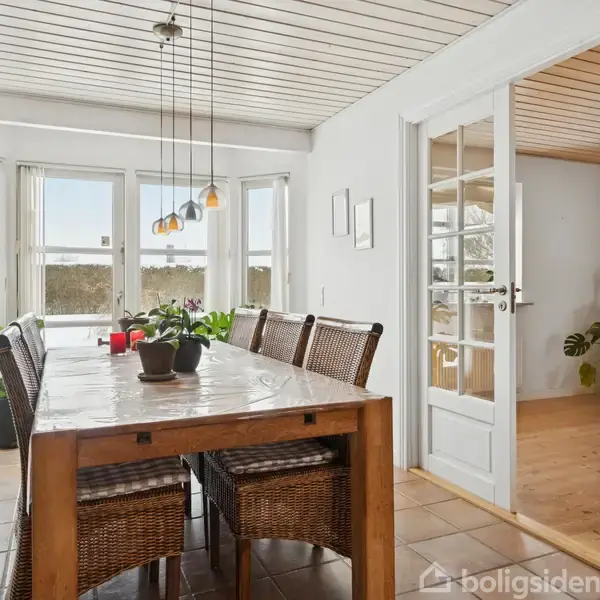 Et spisebord i træ med syv stole omgivet af store vinduer i et lyst rum. Der hænger farvede lamper over bordet, og rummet er åbent til en anden del af huset. Planter står både på og omkring bordet.