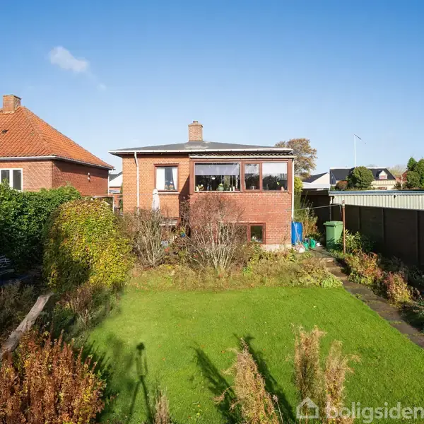 Rødt murstenshus med en stor, velholdt have. Huset har kvistvinduer og ligger mellem to lignende bygninger. Sollys skinner på græsplænen og buske. Klar blå himmel i baggrunden.