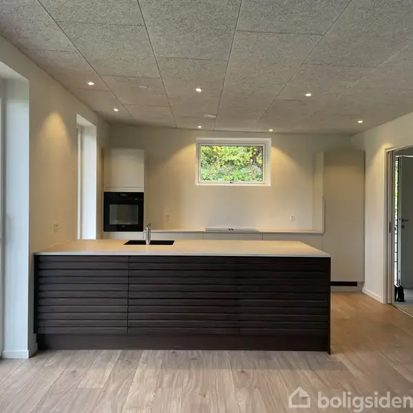 Skab med åbne hylder står langs en hvid væg i et lyst rum med trægulv. Et stort vindue til højre lukker udsigt over grønt område ind.