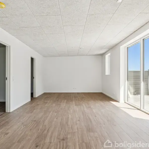 En tom stue med trægulv har to døråbninger til venstre og store vinduer til højre, som giver udsigt til en græsset have.