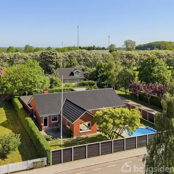 Et rødt murstenshus med mørkt tag står i en velplejet have med pool. Omgivet af grønne træer og nabohuse, på en solrig dag med blå himmel.