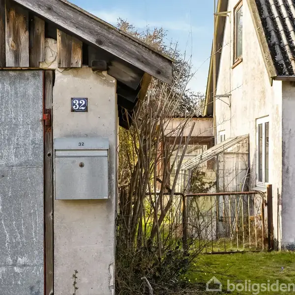 En hvid bygning med en postkasse og husnummer 32 ved siden af en garage. Konteksten er en udendørs gårdscene med en græsplæne og en indgangsport.
