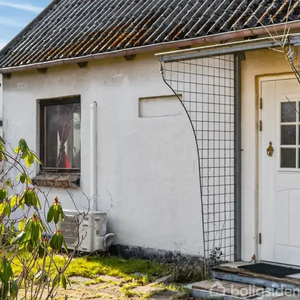 Hvid husfacade med en dør til højre og et lille vindue til venstre. En metalgitterstruktur støtter over døren. Omgivet af nogle grønne buske og flisebelægning.