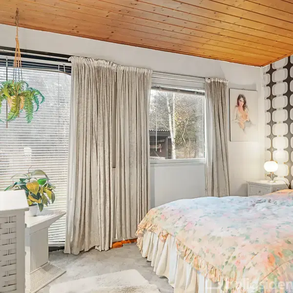 Et soveværelse indeholder en seng med blomstrede sengetøj, omgivet af gardiner og et træloft. Planter hænger ved vinduet, og en lampe står på natbordet ved siden af sengen.