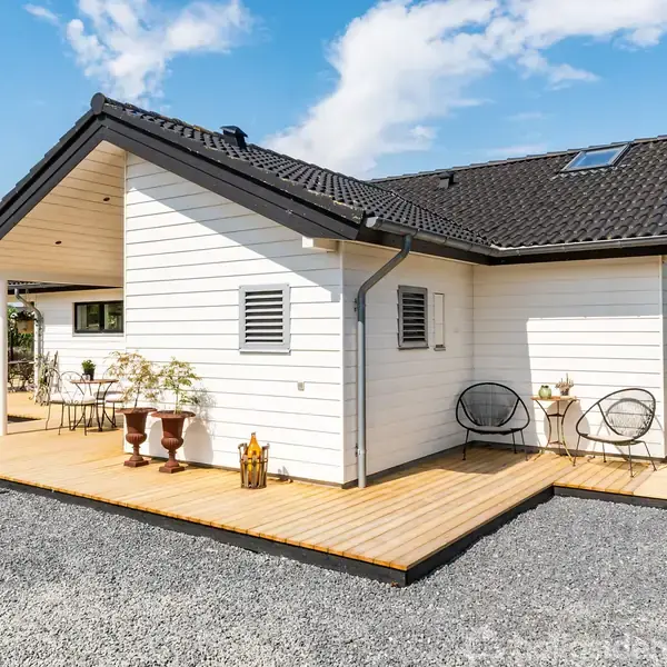 Et hvidt sommerhus står på en stor, grusbelagt grund med en træterrasse. Havemøbler og planter pryder terrassen, omgivet af grønne træer og en blå himmel med skyer.