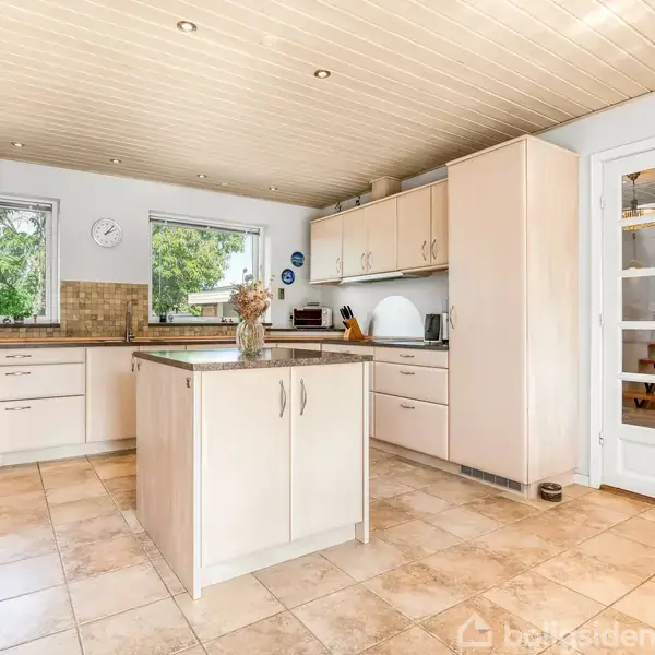 Et lyst køkken med en køkkenø står midt i rummet. Skabe og skuffer omkranser rummet, mens to vinduer giver udsigt til grønne omgivelser. En glasdør leder til næste rum.